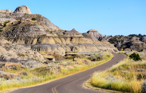 A winding road through rugged, colorful hills under a clear blue sky, surrounded by grass and shrubs.