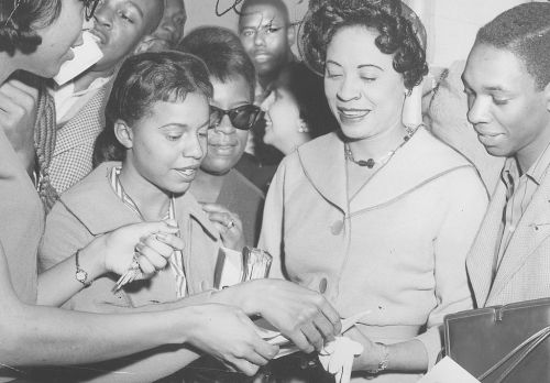 A group of people, including a woman in a light-colored suit, gather around, exchanging papers and smiling.