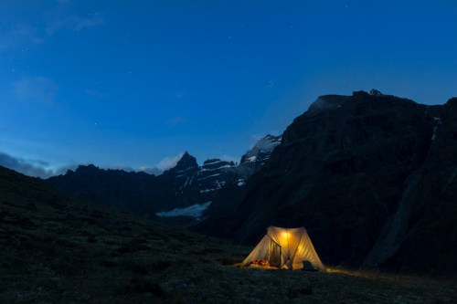 A glowing tent in a mountainous landscape at dusk, with stars visible in the clear blue sky.
