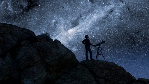 A silhouette of a person with a telescope on a rocky outcrop, gazing at a starry night sky filled with galaxies.
