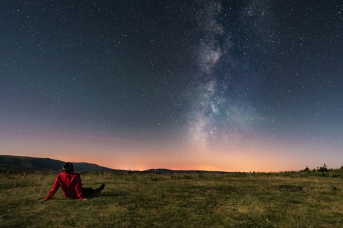 A person in a red jacket sits on grass, gazing at a starry sky with the Milky Way visible above.