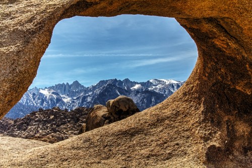 A view of snow-capped mountains through a natural rock arch under a clear blue sky.