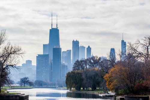 A skyline view of Chicago with tall buildings and a river, surrounded by trees and a cloudy sky.