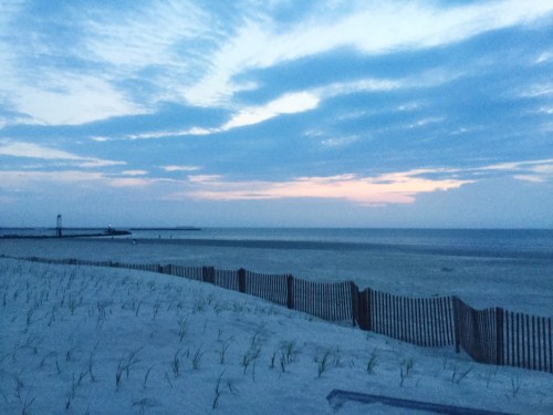 A serene beach at dusk, with gentle waves, a sandy shore, and a wooden fence in the foreground under a cloudy sky.