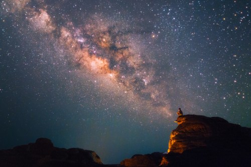 A person sits on a rock under a starry sky, gazing at the Milky Way galaxy above.