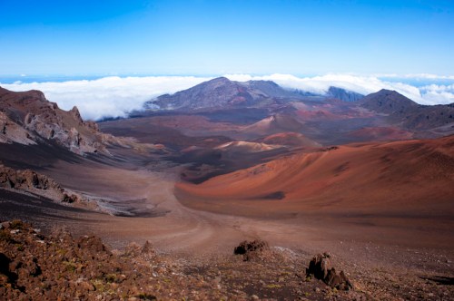 A panoramic view of a volcanic landscape with colorful terrain and clouds in the distance under a clear blue sky.