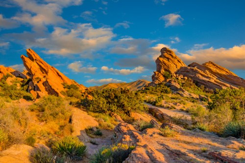 A scenic landscape featuring rocky formations under a blue sky with scattered clouds and desert vegetation.