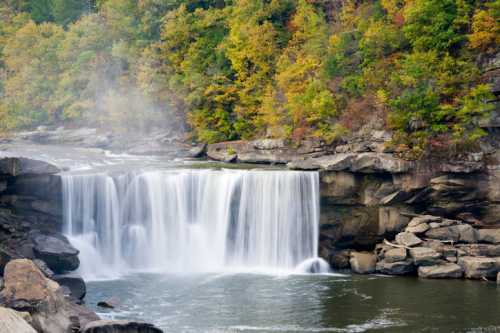 A serene waterfall cascades over rocky cliffs, surrounded by vibrant autumn foliage and mist rising from the water.