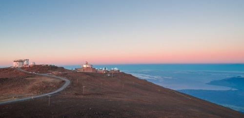 A panoramic view of observatories on a mountain at sunrise, with a clear sky and distant ocean visible.