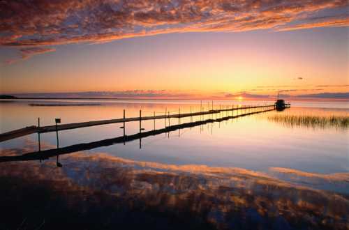 A serene sunset over a calm lake, with a wooden pier extending into the water and colorful clouds reflecting on the surface.