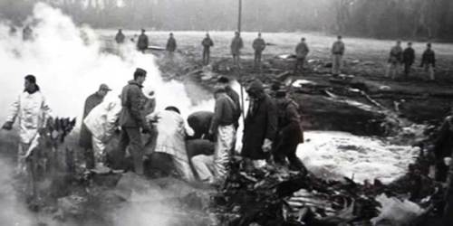 A group of people in protective clothing examines a smoky crash site, with soldiers observing in the background.