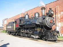 A vintage steam locomotive parked beside a brick building, with smoke billowing from its chimney.