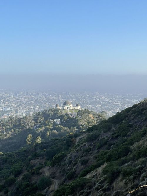 A distant view of a building on a hill, surrounded by greenery, with a hazy city skyline in the background.
