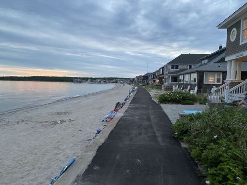 A quiet beachside path lined with houses, leading to a sandy shore under a cloudy sky at dusk.