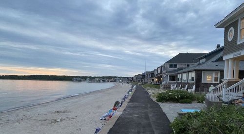 A peaceful beachside scene with a sandy shore, calm water, and houses lining the walkway under a cloudy sky.