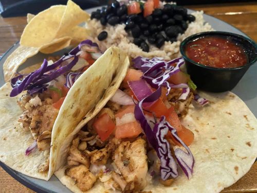 Two tacos filled with grilled chicken, cabbage, and tomatoes, served with rice, black beans, tortilla chips, and salsa.