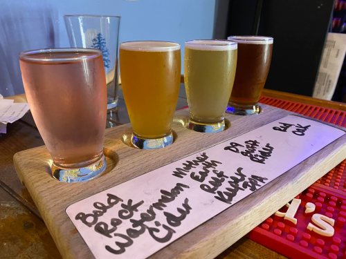 A wooden flight board with four glasses of different craft beers and a cider, labeled with their names.