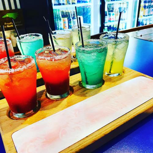 A wooden tray holds five colorful cocktails with straws, featuring vibrant hues of red, green, and yellow.