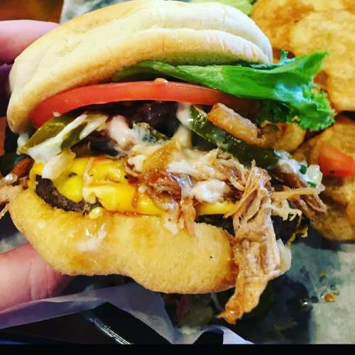 A hand holding a loaded sandwich with pulled pork, cheese, lettuce, tomato, and jalapeños, served with crispy sides.