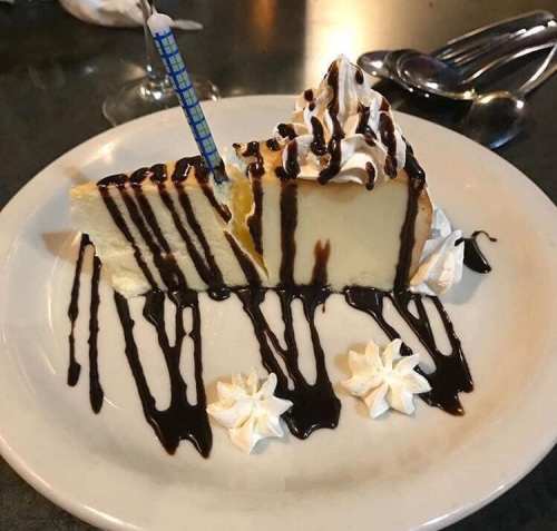 A slice of cheesecake drizzled with chocolate sauce, topped with whipped cream and a birthday candle on a white plate.