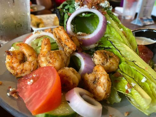 A plate of shrimp salad featuring grilled shrimp, lettuce, tomato, red onion, and a creamy dressing on the side.