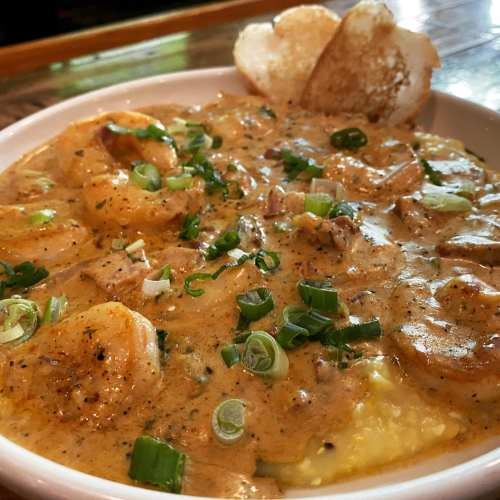 A bowl of creamy shrimp and grits topped with green onions, served with crispy bread on the side.