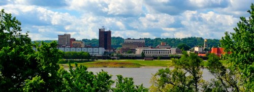 A scenic view of a city skyline across a river, surrounded by greenery and under a partly cloudy sky.