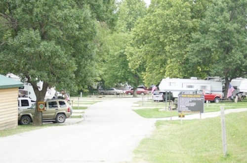 A tree-lined campground with parked RVs and cars along a gravel road, featuring a sign for park regulations.