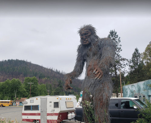 A large, wire sculpture of Bigfoot stands near parked RVs and trees in a rural area.