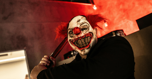 A menacing person in a terrifying clown mask with a bloody mouth and red hair wields a baseball bat.