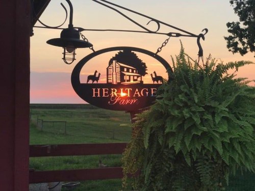 A sign for "Heritage Farm" hangs from a post, surrounded by greenery, with a colorful sunset in the background.