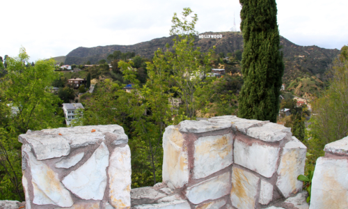View of the Hollywood sign from a stone wall, surrounded by greenery and hills under a cloudy sky.