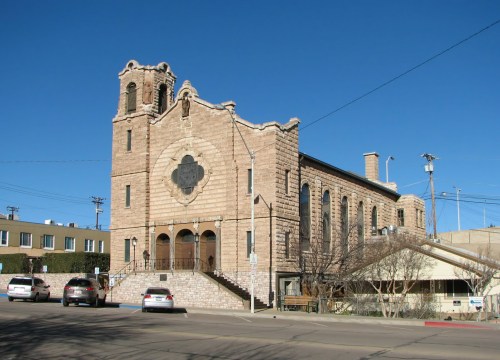 Historic stone church with a bell tower, set against a clear blue sky, surrounded by trees and parked cars.