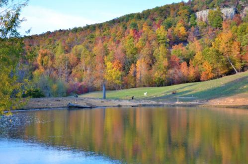 A serene lake reflects vibrant autumn foliage on surrounding hills, with a clear blue sky above.