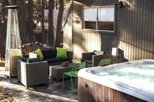 A cozy outdoor seating area with a hot tub, surrounded by trees and a modern cabin. Green cushions add a pop of color.