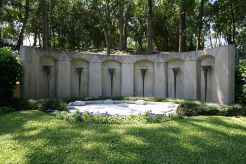 A serene outdoor space featuring a curved stone wall with water features, surrounded by lush greenery and flowers.