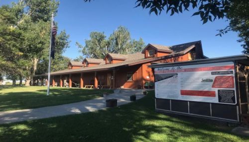 A wooden building with a porch, surrounded by trees and grass, featuring a sign with information in front.