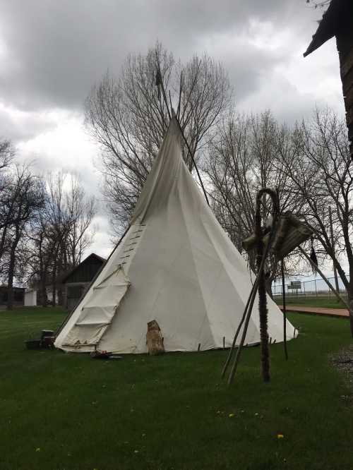 A white teepee stands on green grass, surrounded by trees under a cloudy sky.