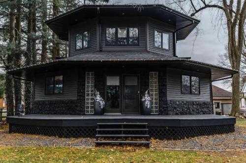 A modern black house with a unique octagonal shape, featuring large windows and a front porch surrounded by trees.