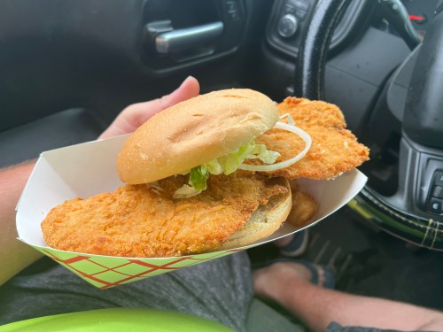 A hand holding a large fried chicken sandwich with lettuce and onion, inside a takeout container in a car.