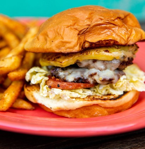 A juicy burger with melted cheese, lettuce, tomato, and grilled pineapple, served with crispy fries on a pink plate.