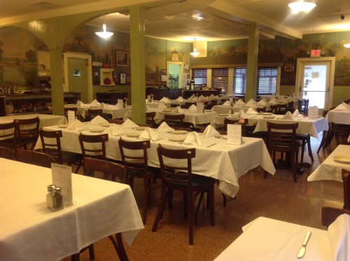 A spacious restaurant with neatly arranged tables, white tablecloths, and a mural on the wall.