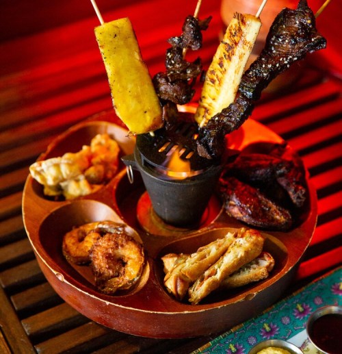 A wooden platter with assorted grilled skewers, shrimp, and fried snacks, set against a warm, colorful background.