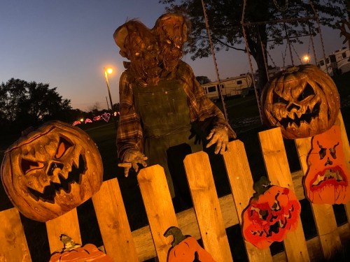 A spooky scarecrow with a grotesque mask stands by carved pumpkins at dusk, surrounded by a wooden fence.