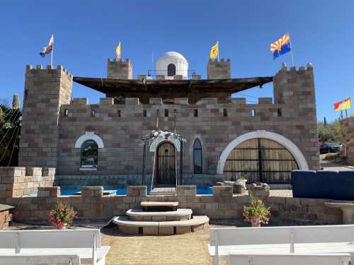 A stone castle with flags, featuring a round tower and a pool in front, set against a clear blue sky.