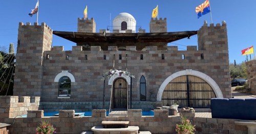 A whimsical stone castle with a dome, flags, and a pool in front, set against a clear blue sky.