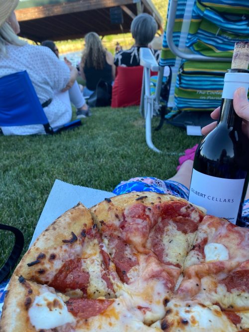 A hand holds a bottle of wine and a slice of pizza, with people sitting in the background at an outdoor event.