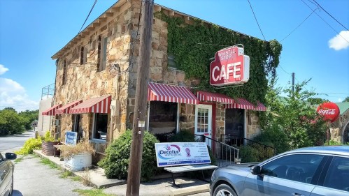 A rustic stone building with a red and white striped awning, featuring a sign for Whistle Stop Café and nearby signage.