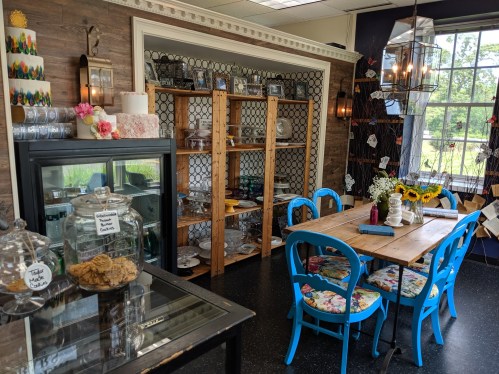 A cozy café interior with a wooden table, blue chairs, shelves of dishes, and a display of baked goods. Sunflowers on the table.