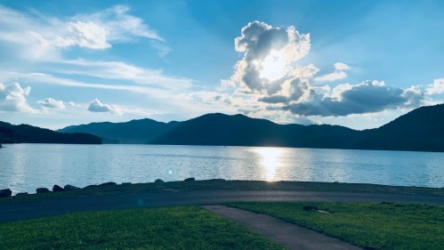 A serene lake scene with mountains in the background, under a bright sky with clouds and sunlight reflecting on the water.